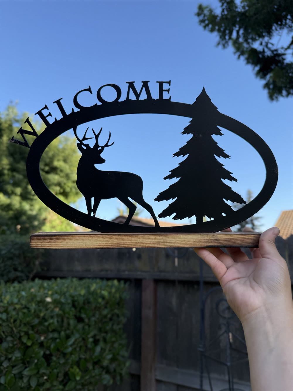 A steel Welcome sign with a buck and a pine tree inside an oval, on a wood base.